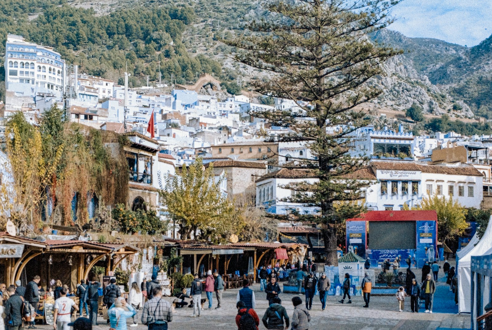 People gather in a bustling town square with blue buildings.